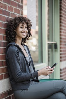 Side view of cheerful young female millennial with curly hair in activewear sitting on bench near brick building and smiling while talking on smartphone via TWS earphones