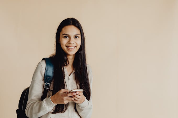 Teen girl with backpack using a smartphone against a beige background, smiling.
